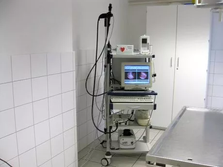 Medizinisches Gerät mit Monitor und Tastatur auf einem Rollwagen in einem weißen, gefliesten Raum neben einem Untersuchungstisch