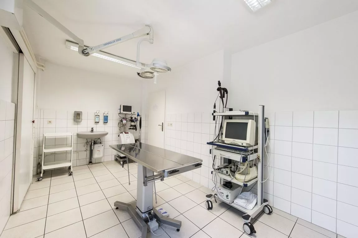Steriler Behandlungsraum mit OP-Tisch, medizinischem Monitor auf Rollwagen, Waschbecken und weißen Fliesen an Wand und Boden