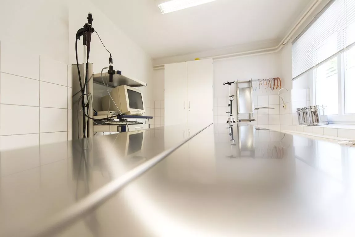 Leerer, heller Behandlungsraum mit langem, glänzendem Metalltisch, medizinischem Gerät und Monitor an der Wand