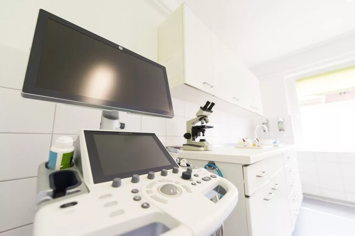 Ultraschallgerät mit Monitor und Bedienelementen in einem hellen Untersuchungsraum mit Mikroskop und Spüle im Hintergrund