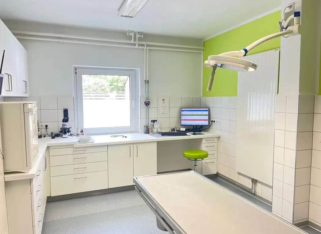Behandlungsraum mit Untersuchungsliege, grünem Hocker, Monitor, Hängeschränken und großem Fenster